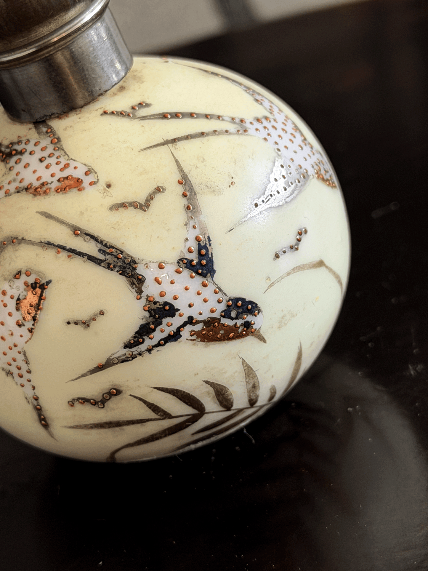 A close-up of a decorative spherical bottle or vase with a cream-colored body, hand-painted birds and leaves in black, white, and copper-gold, and a silver-toned metal cap.