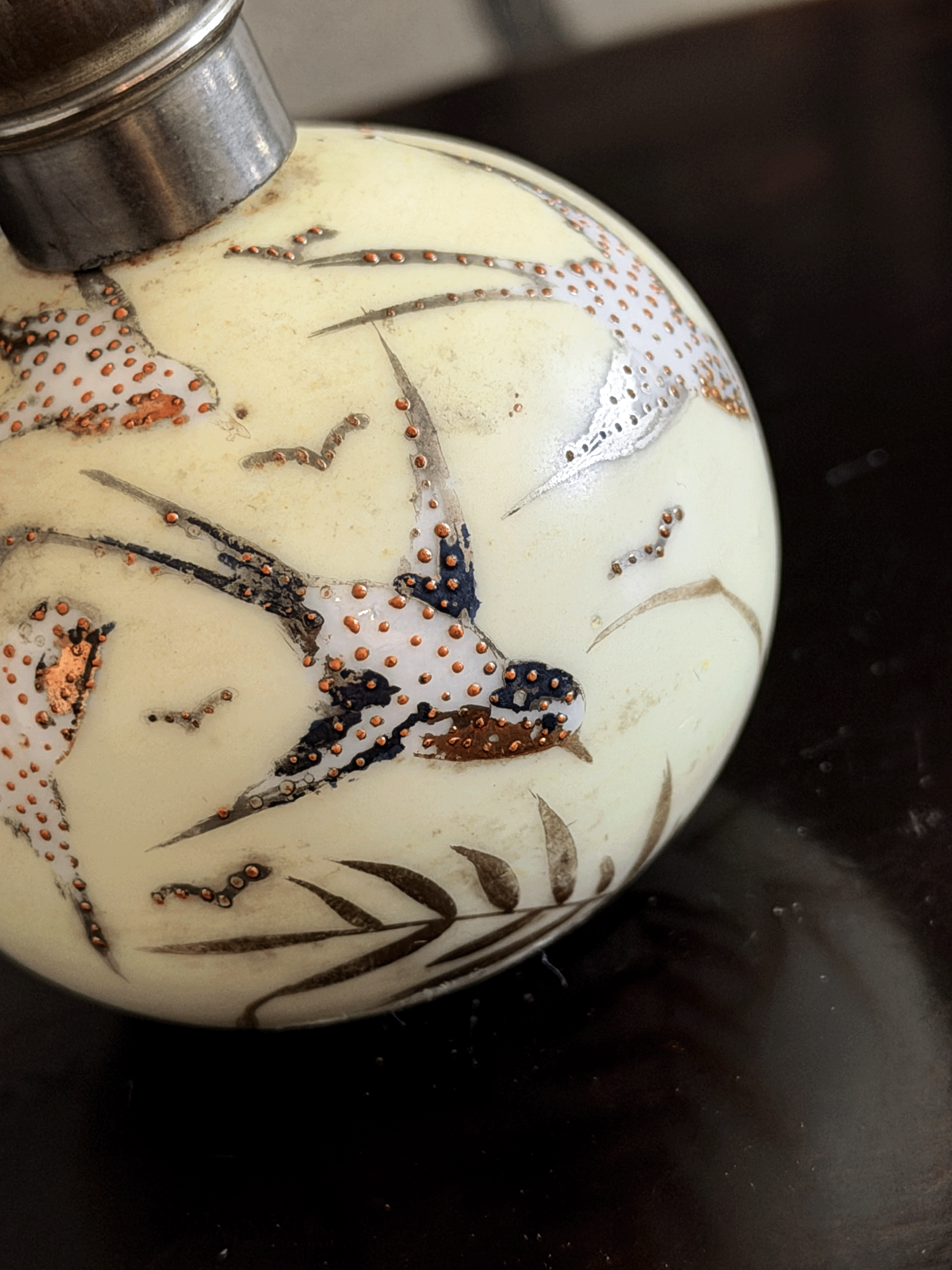 A close-up of a decorative spherical bottle or vase with a cream-colored body, hand-painted birds and leaves in black, white, and copper-gold, and a silver-toned metal cap.