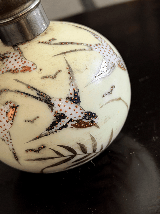 A close-up of a decorative spherical bottle or vase with a cream-colored body, hand-painted birds and leaves in black, white, and copper-gold, and a silver-toned metal cap.