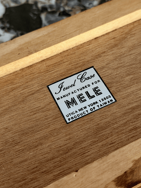 A close-up of a wooden jewelry box with a white label that reads 'Jewel Case Manufactured for MELE Utica New York' and 'Product of Taiwan'.