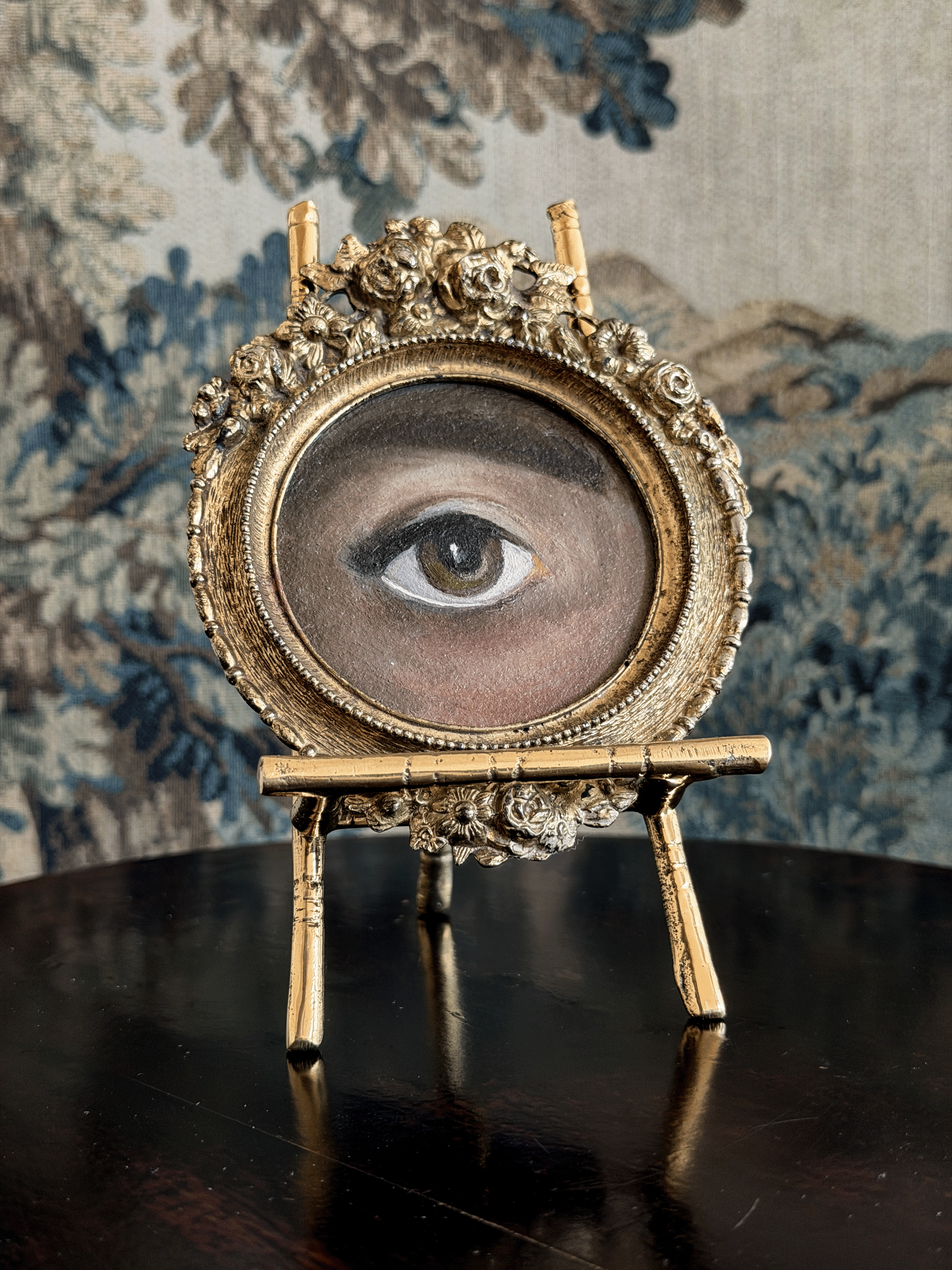 An ornate gold-framed oval tabletop mirror or picture frame featuring a painted human eye, displayed on a small gold stand over a dark reflective surface against a blue floral tapestry background.