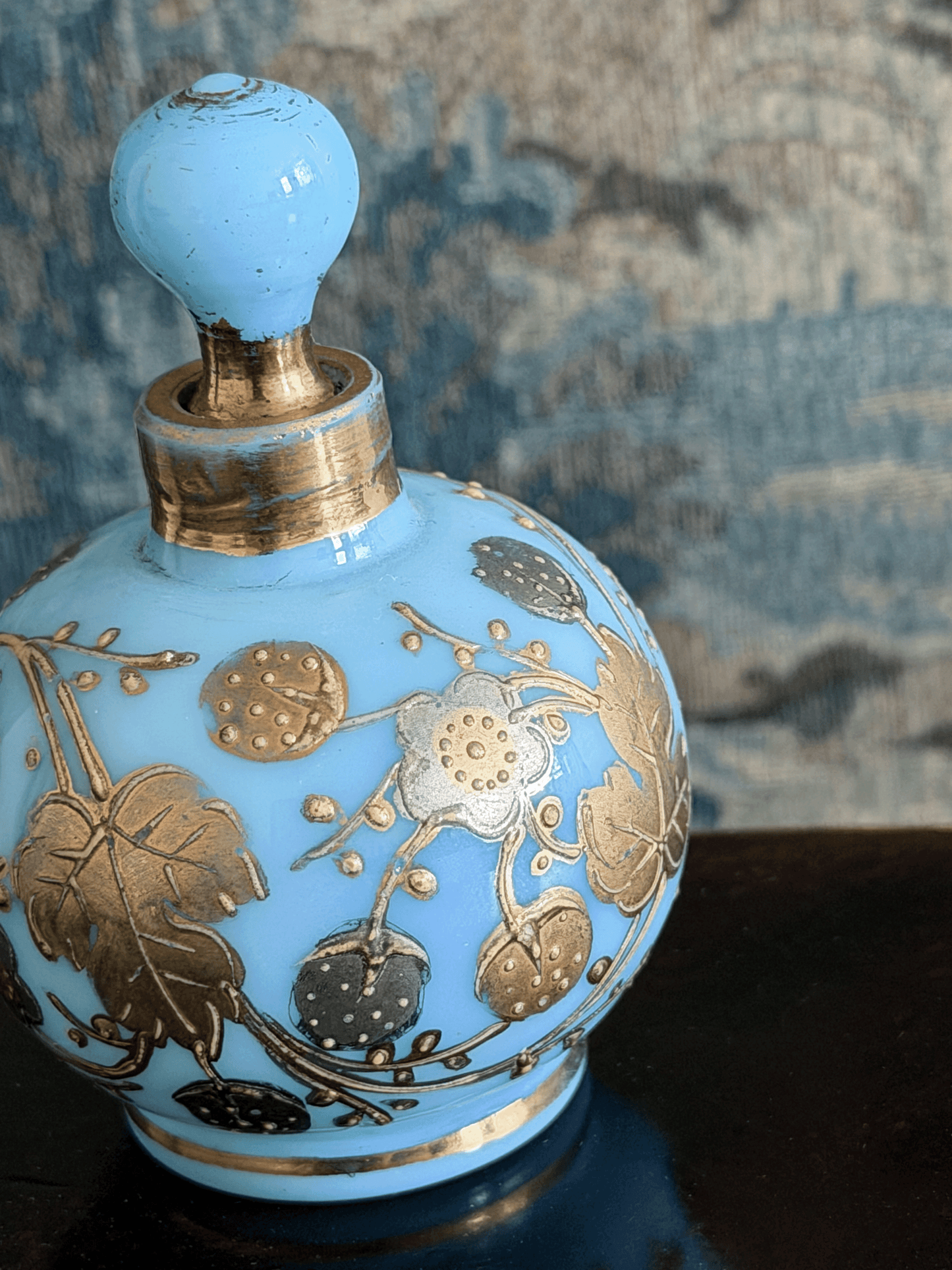 A light blue glass perfume bottle with a matching stopper, decorated with metallic gold floral motifs, set against a textured blue-gray background.