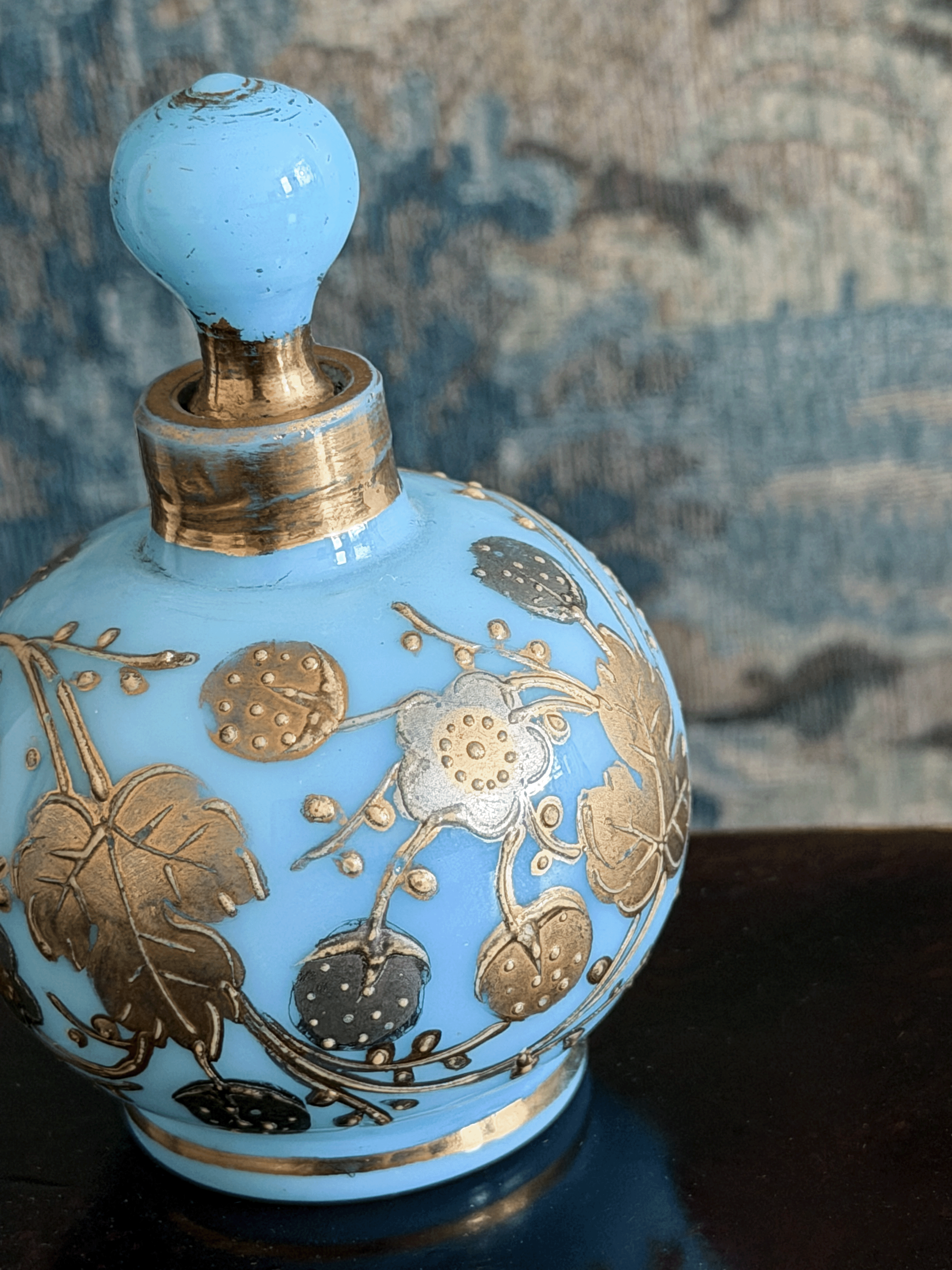 A light blue glass perfume bottle with a matching stopper, decorated with metallic gold floral motifs, set against a textured blue-gray background.