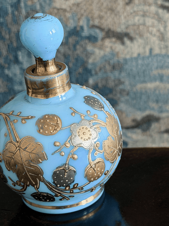 A light blue glass perfume bottle with a matching stopper, decorated with metallic gold floral motifs, set against a textured blue-gray background.