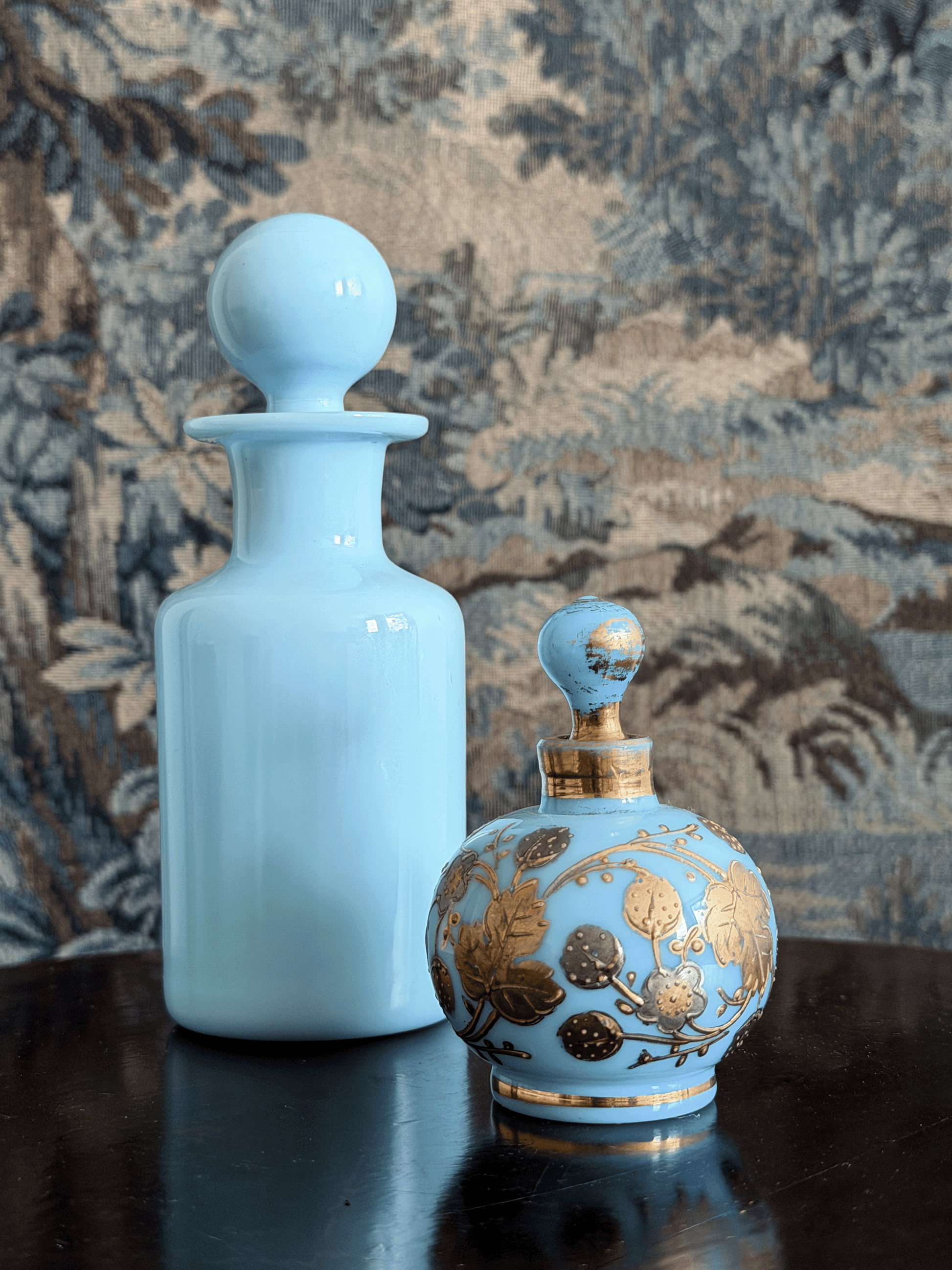 A light blue glass perfume bottle with a round stopper and a smaller ornate perfume atomizer with gold floral detailing, set on a dark reflective surface against a patterned tapestry background.