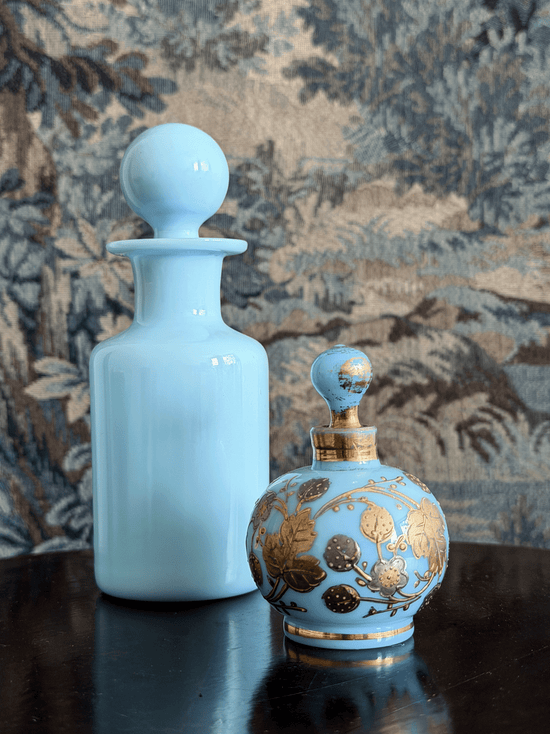 A light blue glass perfume bottle with a round stopper and a smaller ornate perfume atomizer with gold floral detailing, set on a dark reflective surface against a patterned tapestry background.