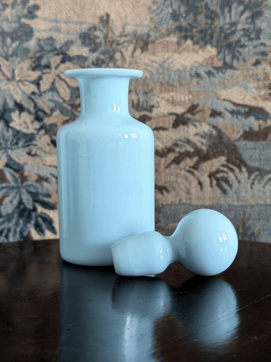A light blue opaline glass bottle with a matching small bulb-shaped stopper, displayed on a dark reflective surface against a patterned tapestry background.