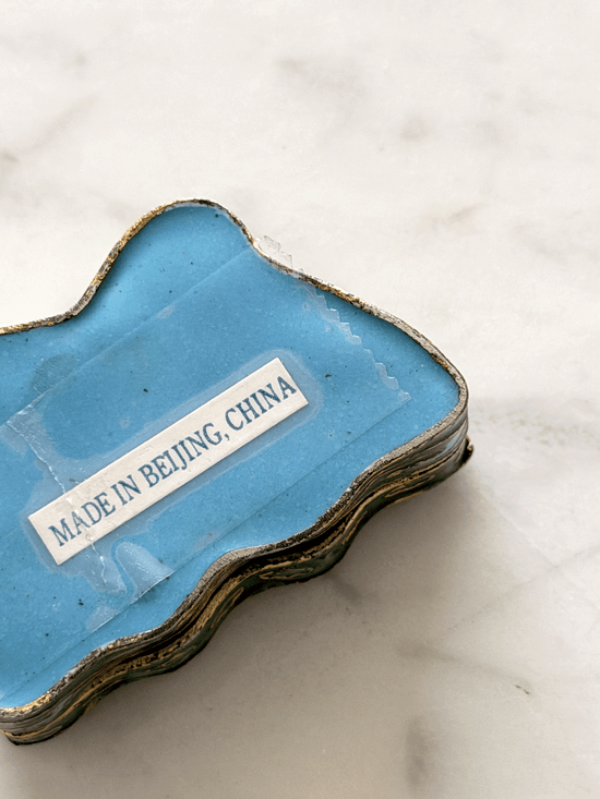 A small butterfly-shaped blue enamel jewelry box with a gold-toned edge, photographed on a light marble surface. A small white label  reads 'MADE IN BEIJING, CHINA'.