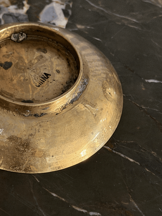 A close-up of a vintage brass dish base, showing a tarnished gold finish with dark patina and a stamped 'CHINA' mark, resting on a dark marble surface.