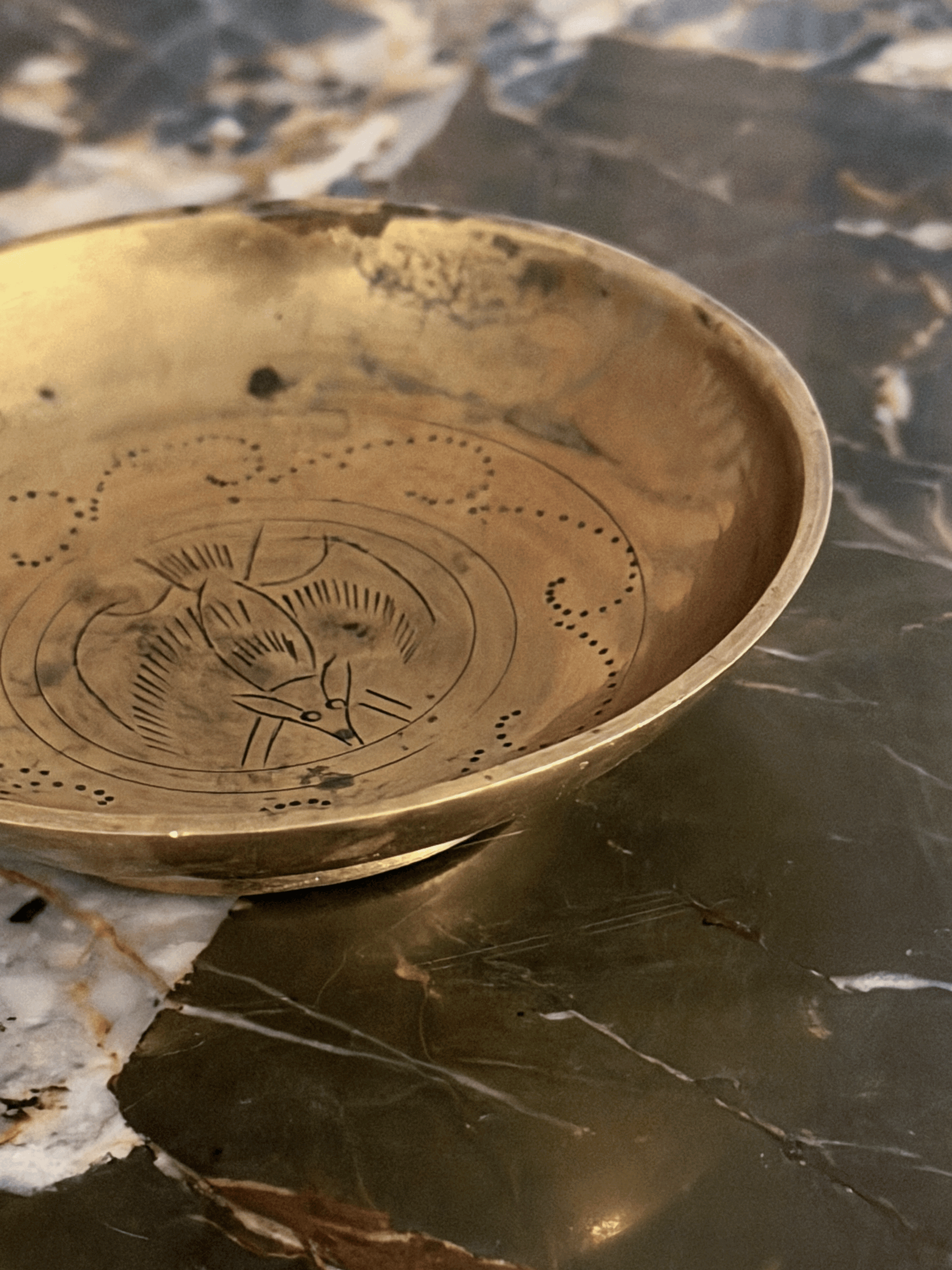A round brass plate with engraved lucky fu bat and dotted patterns, resting on a polished dark marble surface.