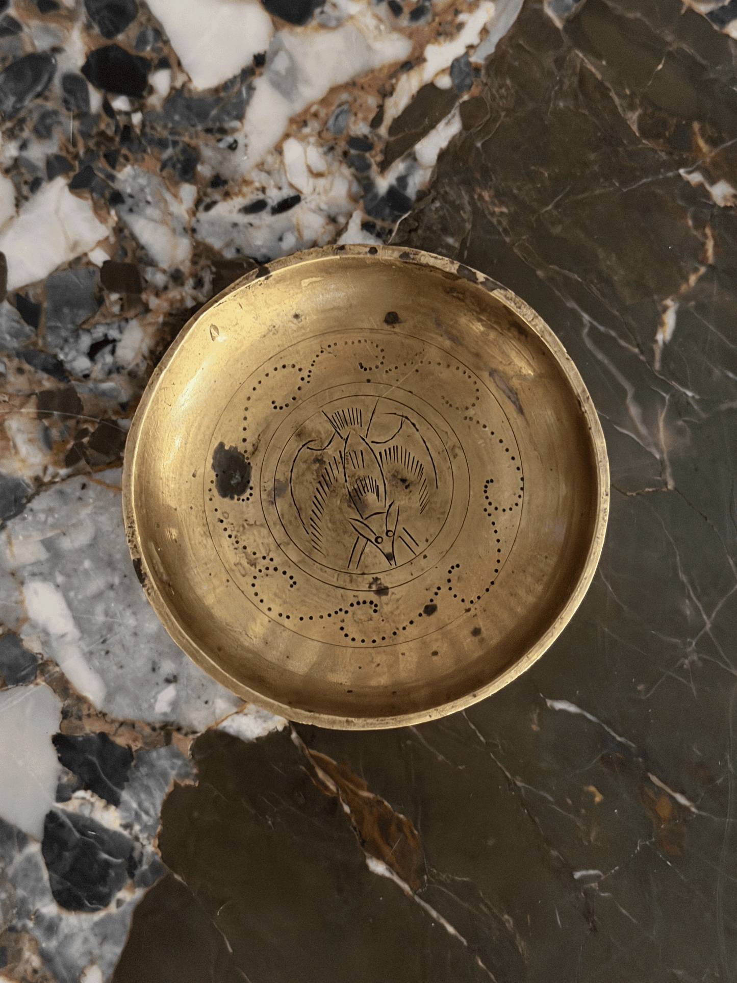 A round brass trinket dish with engraved bat and patterning, photographed from above on a dark and light marbled stone surface.