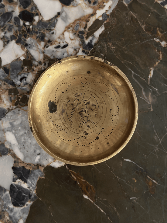 A round brass trinket dish with engraved bat and patterning, photographed from above on a dark and light marbled stone surface.