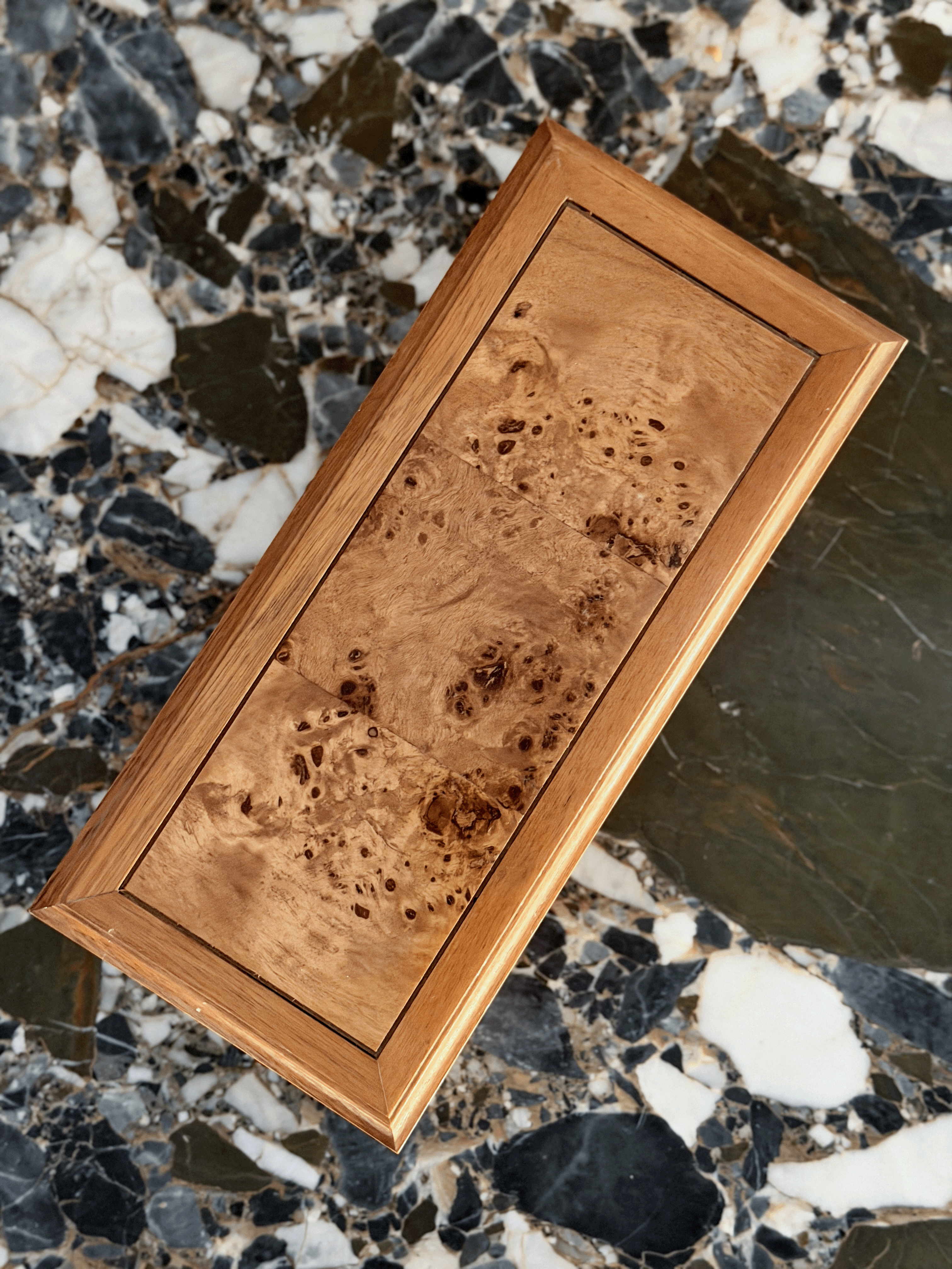 A rectangular wooden box with a rich burl wood grain pattern and a polished beveled frame, shown diagonally on a marble surface.