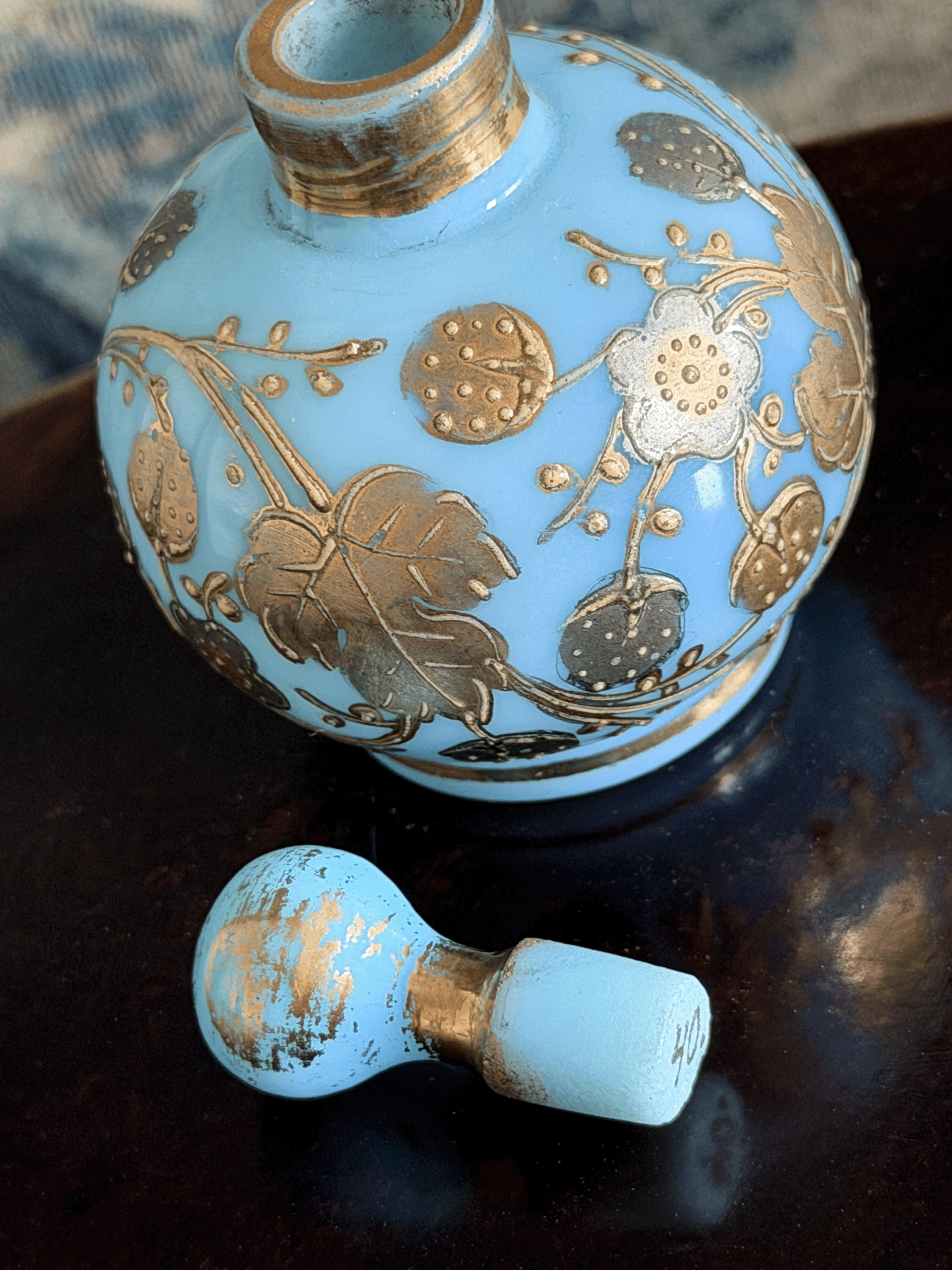 A light blue opaline glass perfume bottle with gold floral patterns, shown with its matching light blue and gold cap resting on a dark reflective surface.