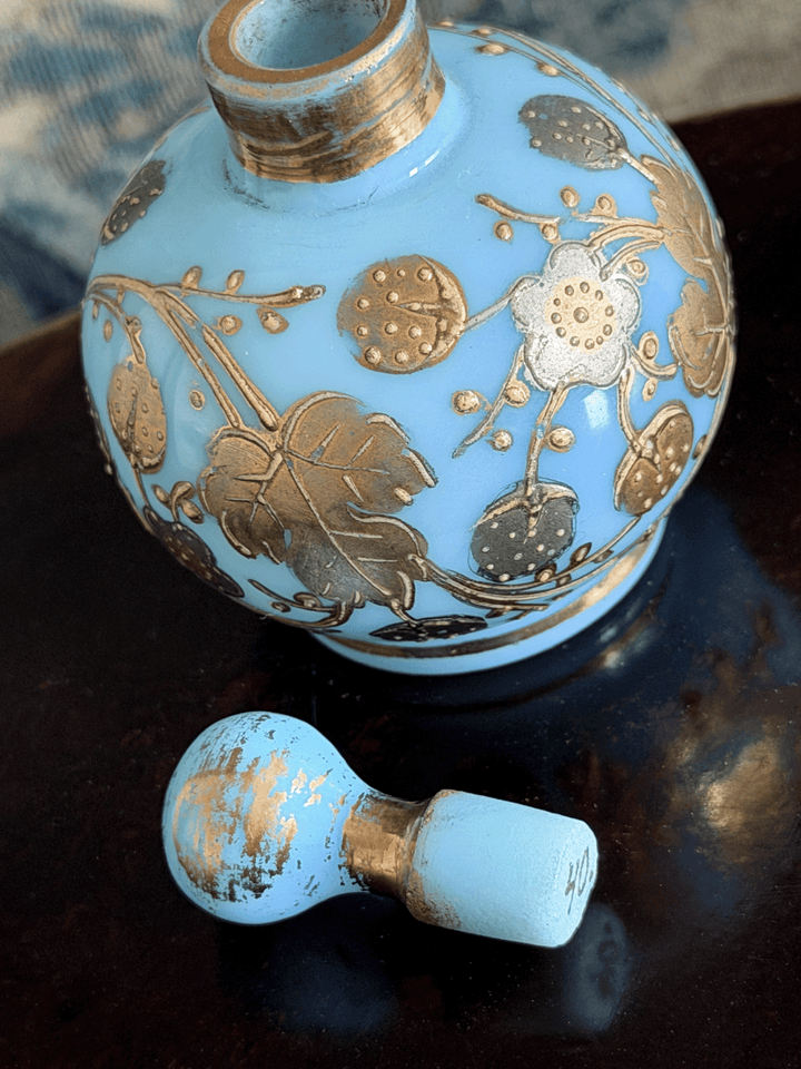 A light blue opaline glass perfume bottle with gold floral patterns, shown with its matching light blue and gold cap resting on a dark reflective surface.