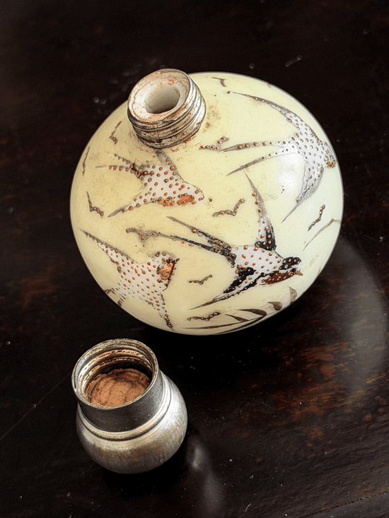 An antique ceramic bottle with a cream-colored body decorated with brown and red spotted bird motifs, shown with its metal cap removed on a dark wooden surface.