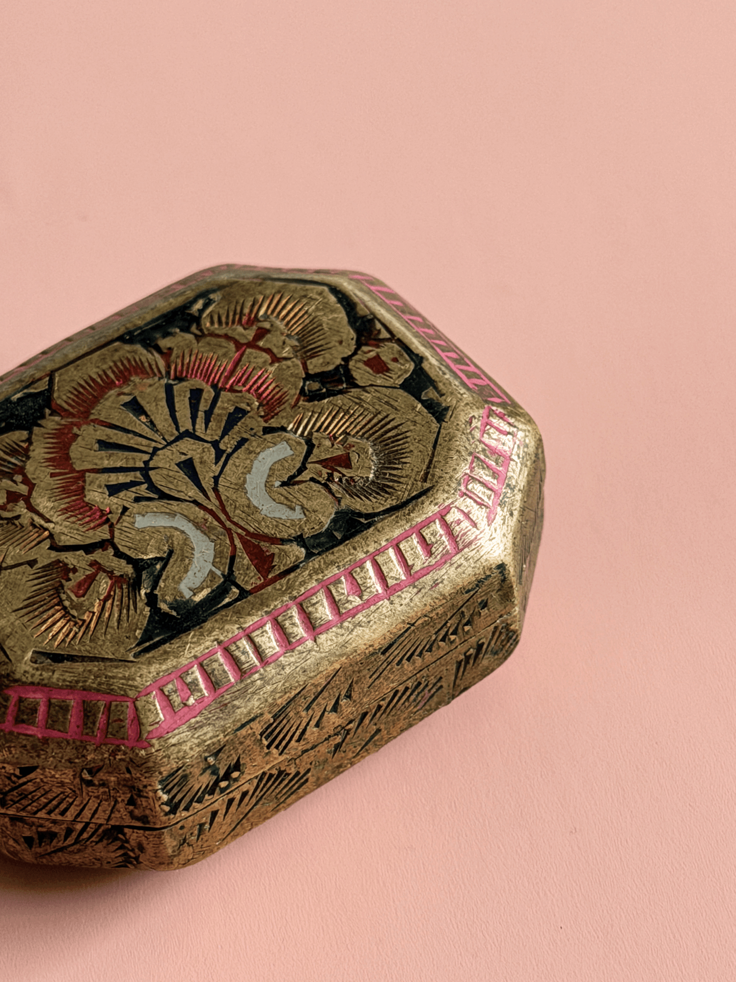 A close-up of an ornate hexagonal decorative box with a gold-toned metallic finish, hand-painted floral motifs in red, black, and cream, and a pink geometric border, set against a solid pink background.