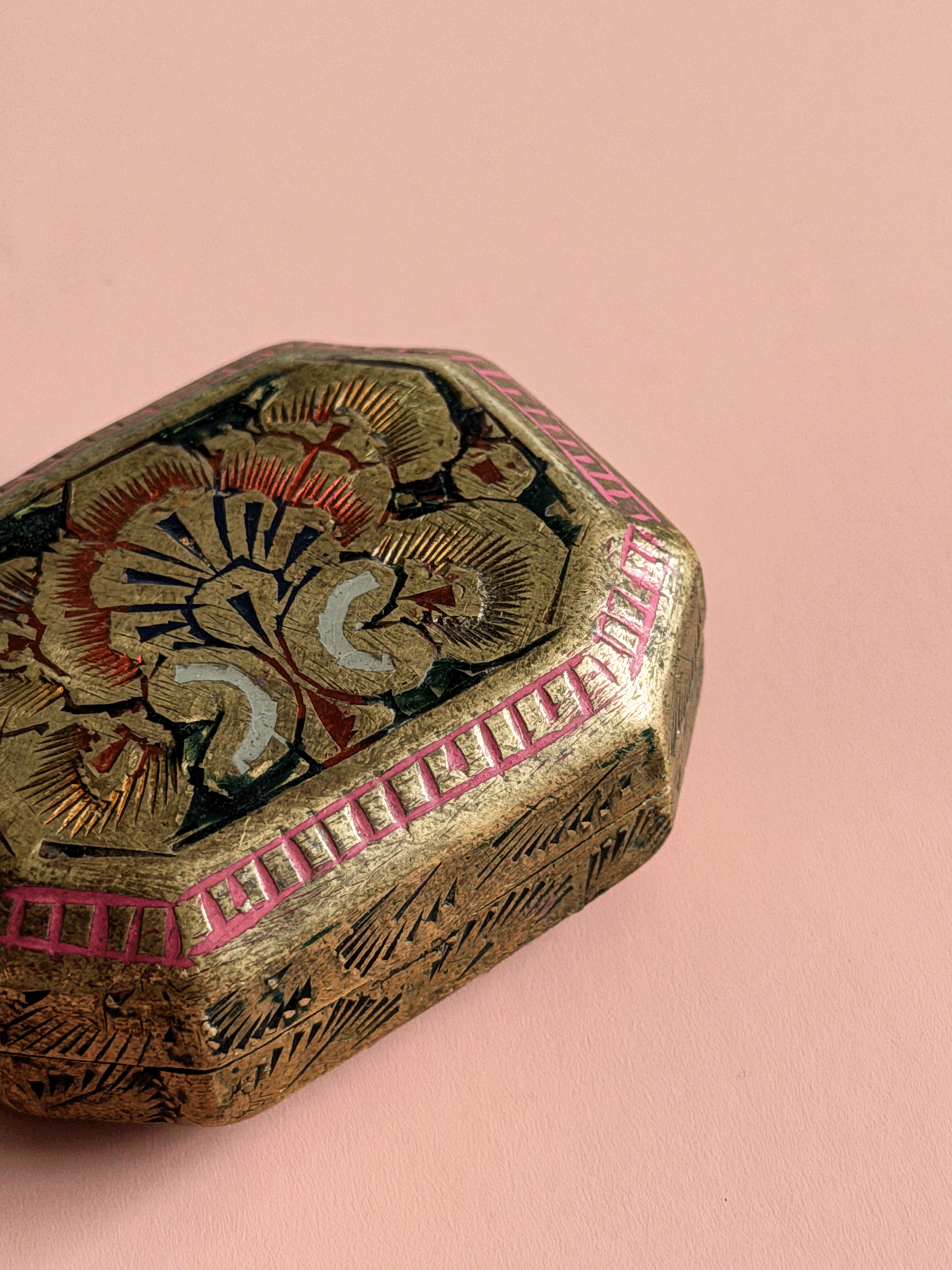 A close-up of an ornate hexagonal decorative box with a gold-toned metallic finish, hand-painted floral motifs in red, black, and cream, and a pink geometric border, set against a solid pink background.