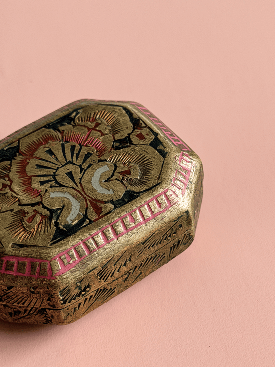 A close-up of an ornate hexagonal decorative box with a gold-toned metallic finish, hand-painted floral motifs in red, black, and cream, and a pink geometric border, set against a solid pink background.