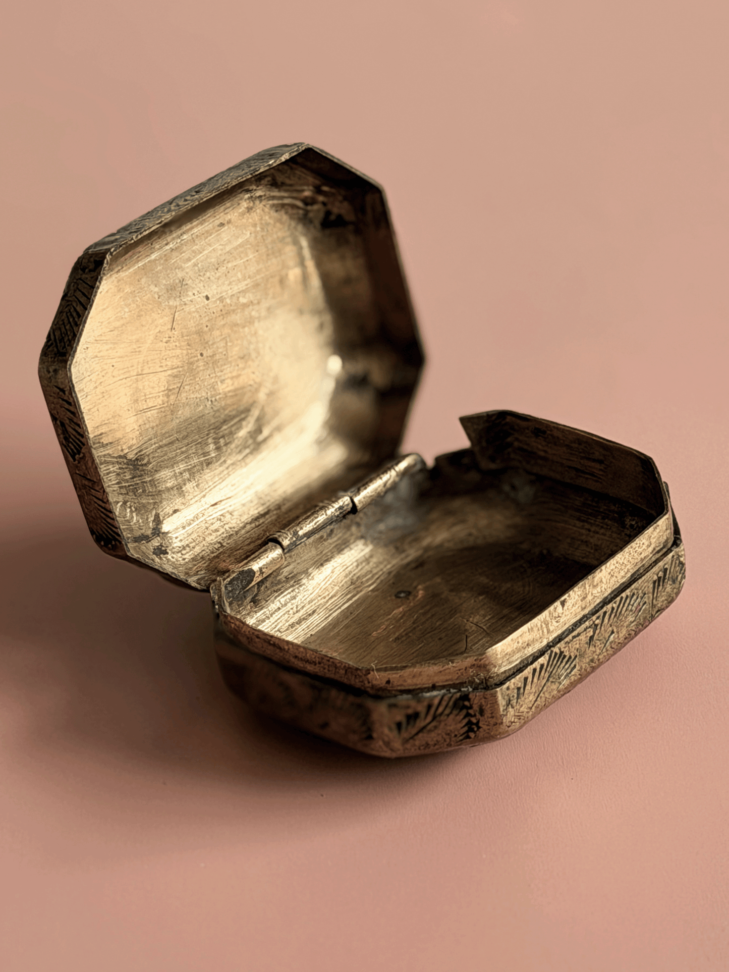 An antique octagonal metal box with an open lid, showing a tarnished brass or bronze finish, set against a solid dusty pink background.