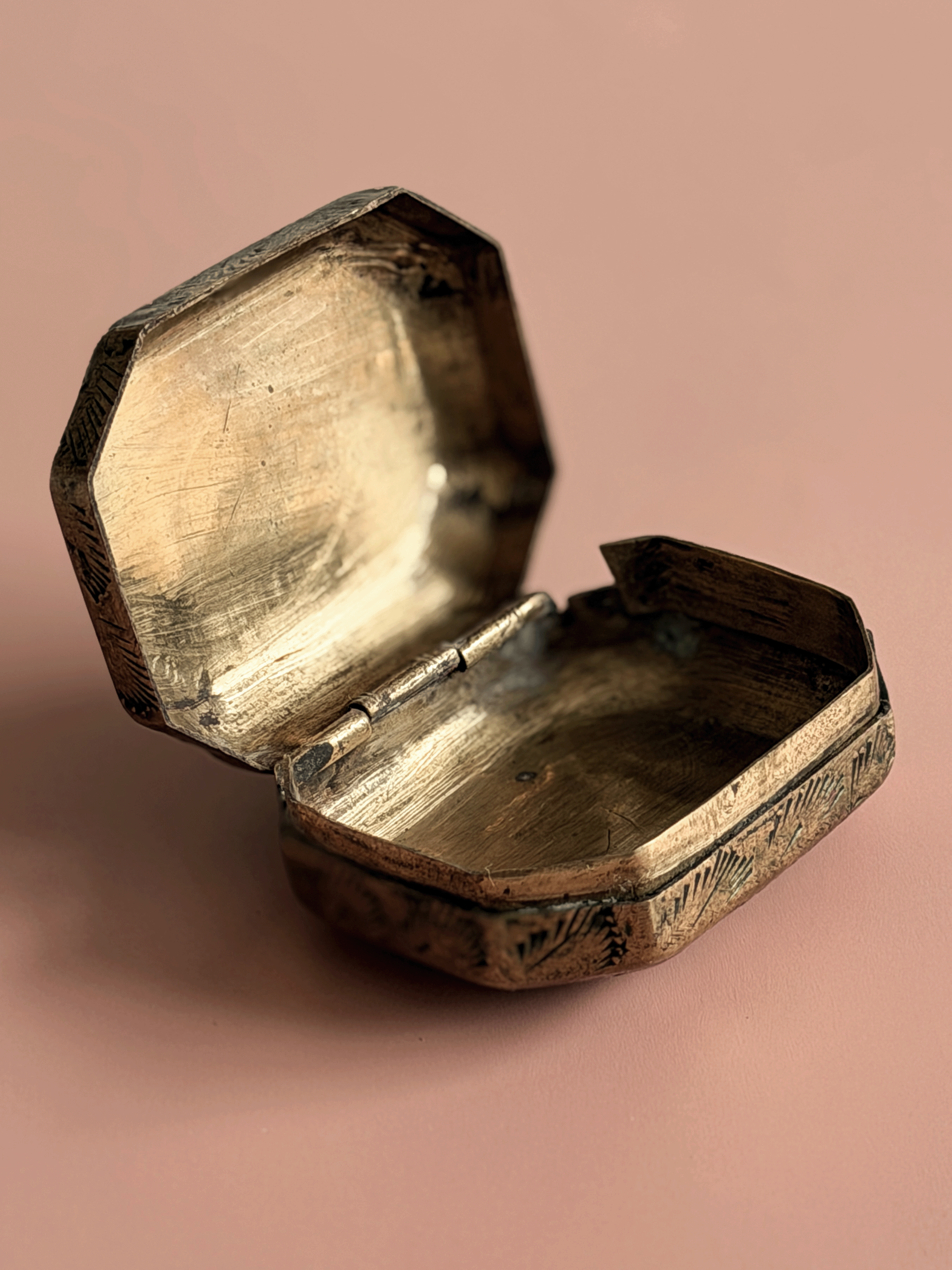 An antique octagonal metal box with an open lid, showing a tarnished brass or bronze finish, set against a solid dusty pink background.