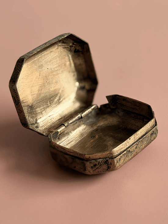 An antique octagonal metal box with an open lid, showing a tarnished brass or bronze finish, set against a solid dusty pink background.