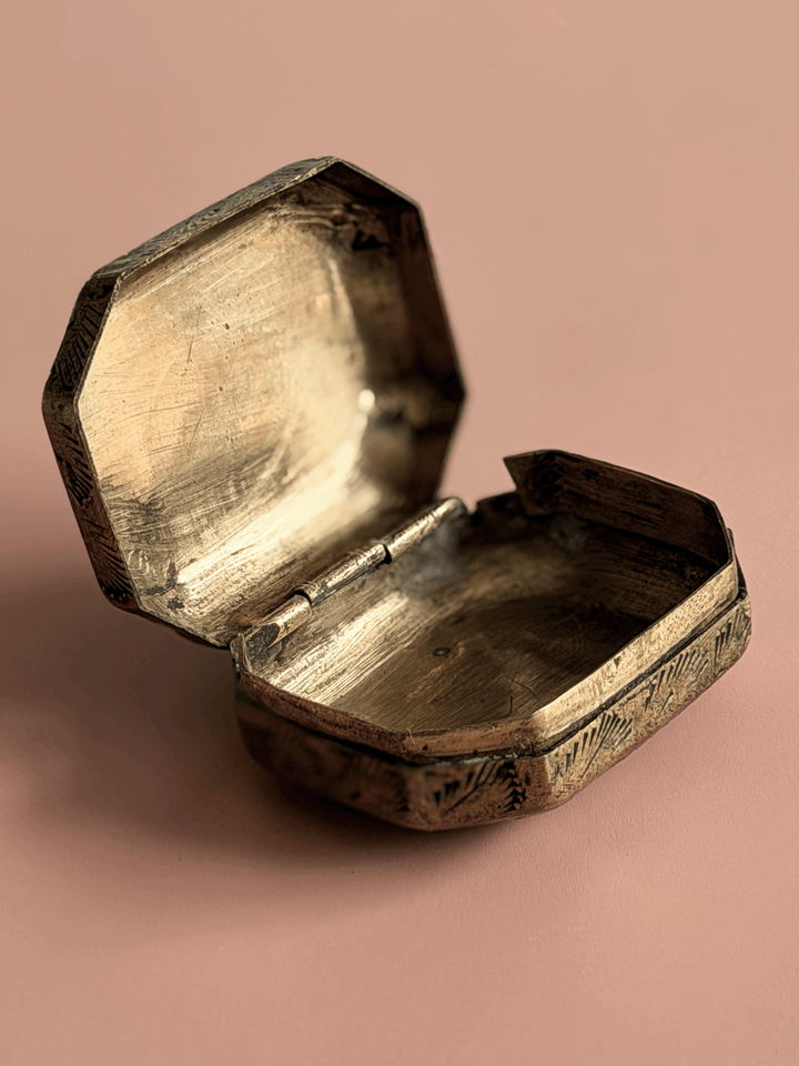An antique octagonal metal box with an open lid, showing a tarnished brass or bronze finish, set against a solid dusty pink background.