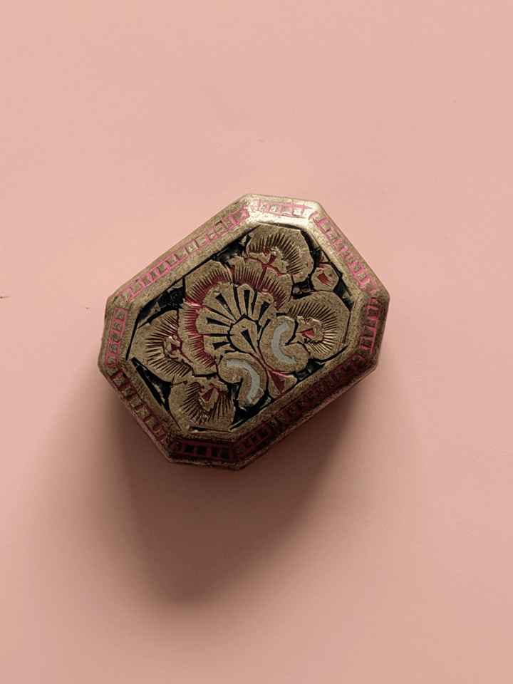 An overhead view of a small, antique octagonal brass trinket box against a pink background. The lid features an intricate, hand-engraved floral design filled with black enamel, surrounded by a border of bright pink enameled geometric cells.