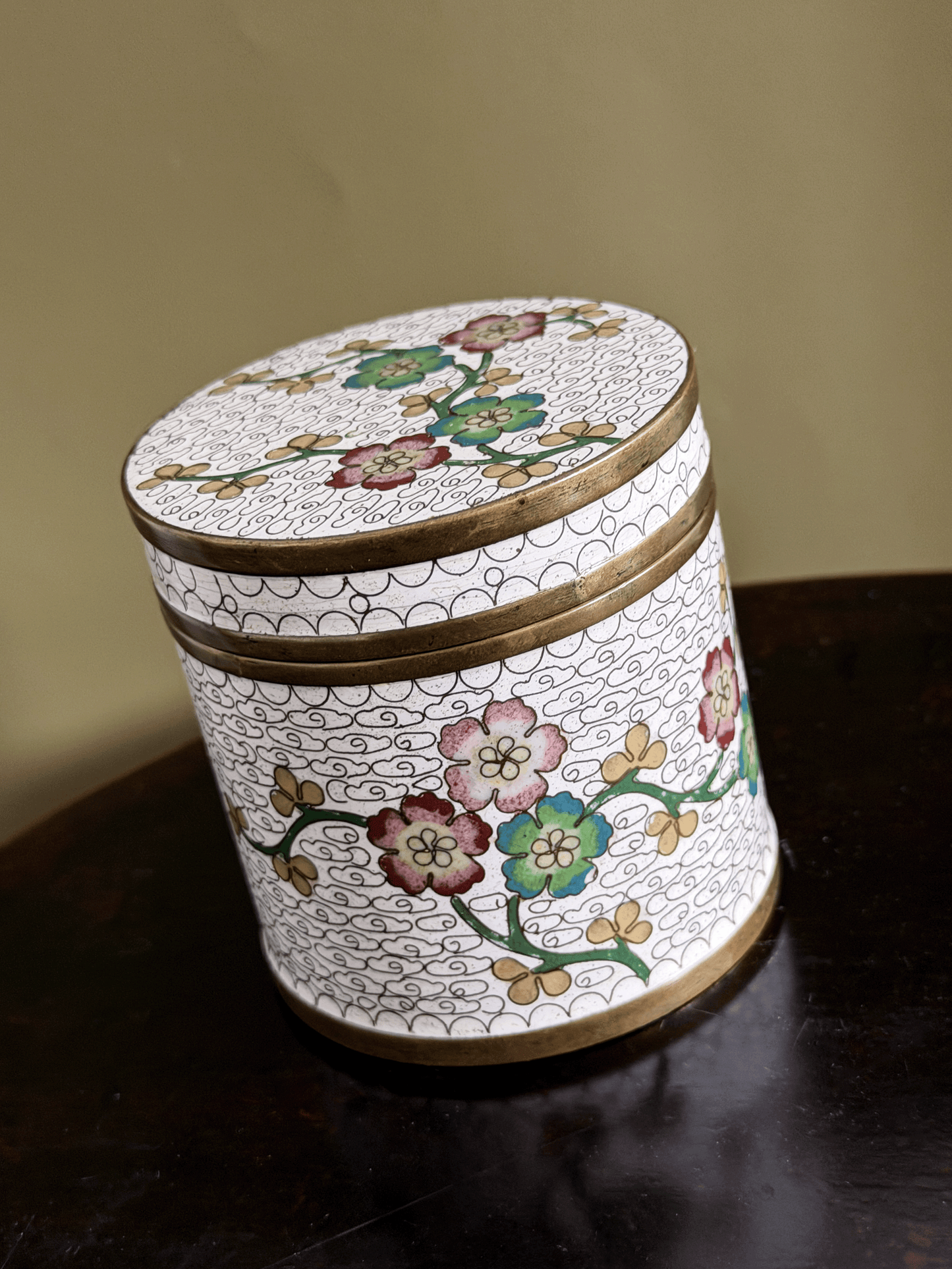 A round decorative lidded box with a white enamel-style background, hand-painted floral motifs in pink, green, and gold, and gold-toned metal trim, shown on a dark wooden surface against a beige wall.