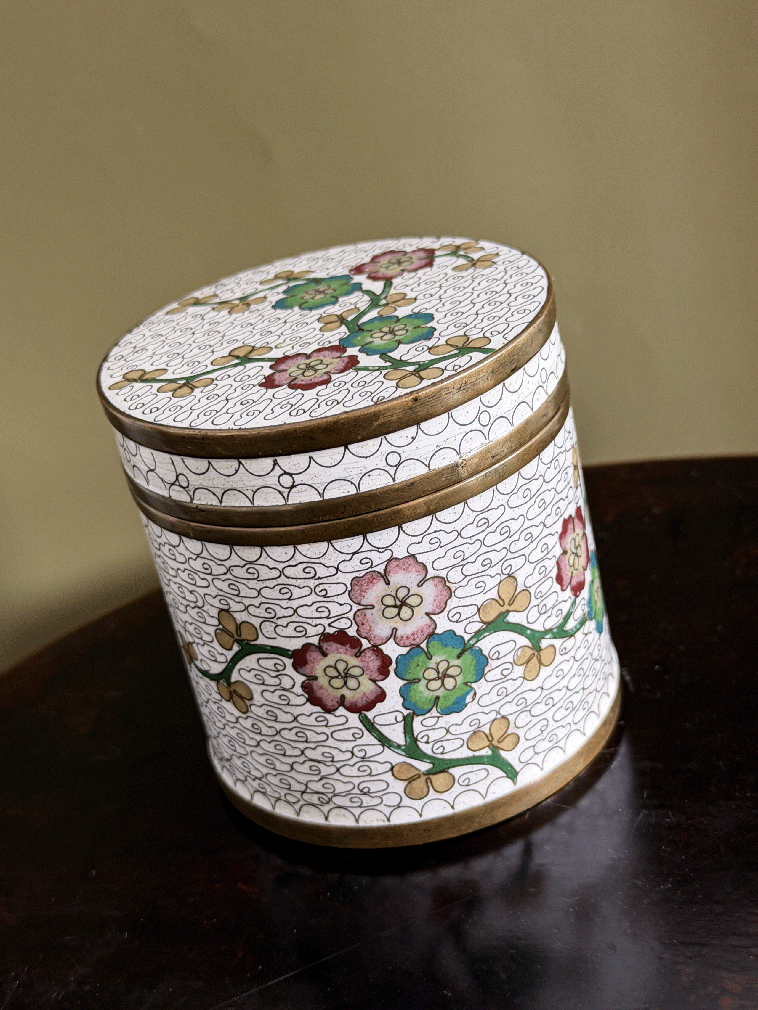 A round decorative lidded box with a white enamel-style background, hand-painted floral motifs in pink, green, and gold, and gold-toned metal trim, shown on a dark wooden surface against a beige wall.