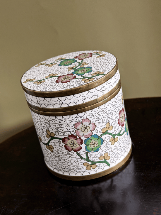 A round decorative lidded box with a white enamel-style background, hand-painted floral motifs in pink, green, and gold, and gold-toned metal trim, shown on a dark wooden surface against a beige wall.