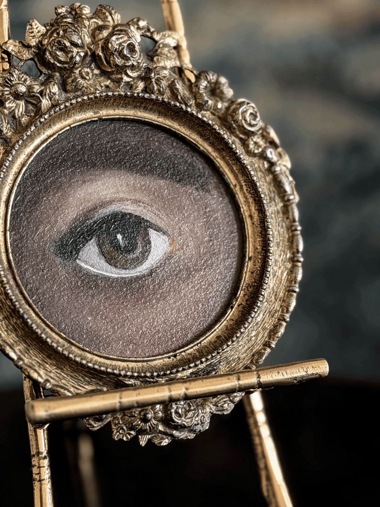 An ornate antique-style gold picture frame holding a painted human eye, displayed on a small stand against a dark blurred background.