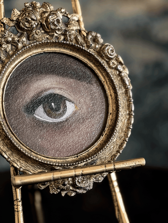 An ornate antique-style gold picture frame holding a painted human eye, displayed on a small stand against a dark blurred background.