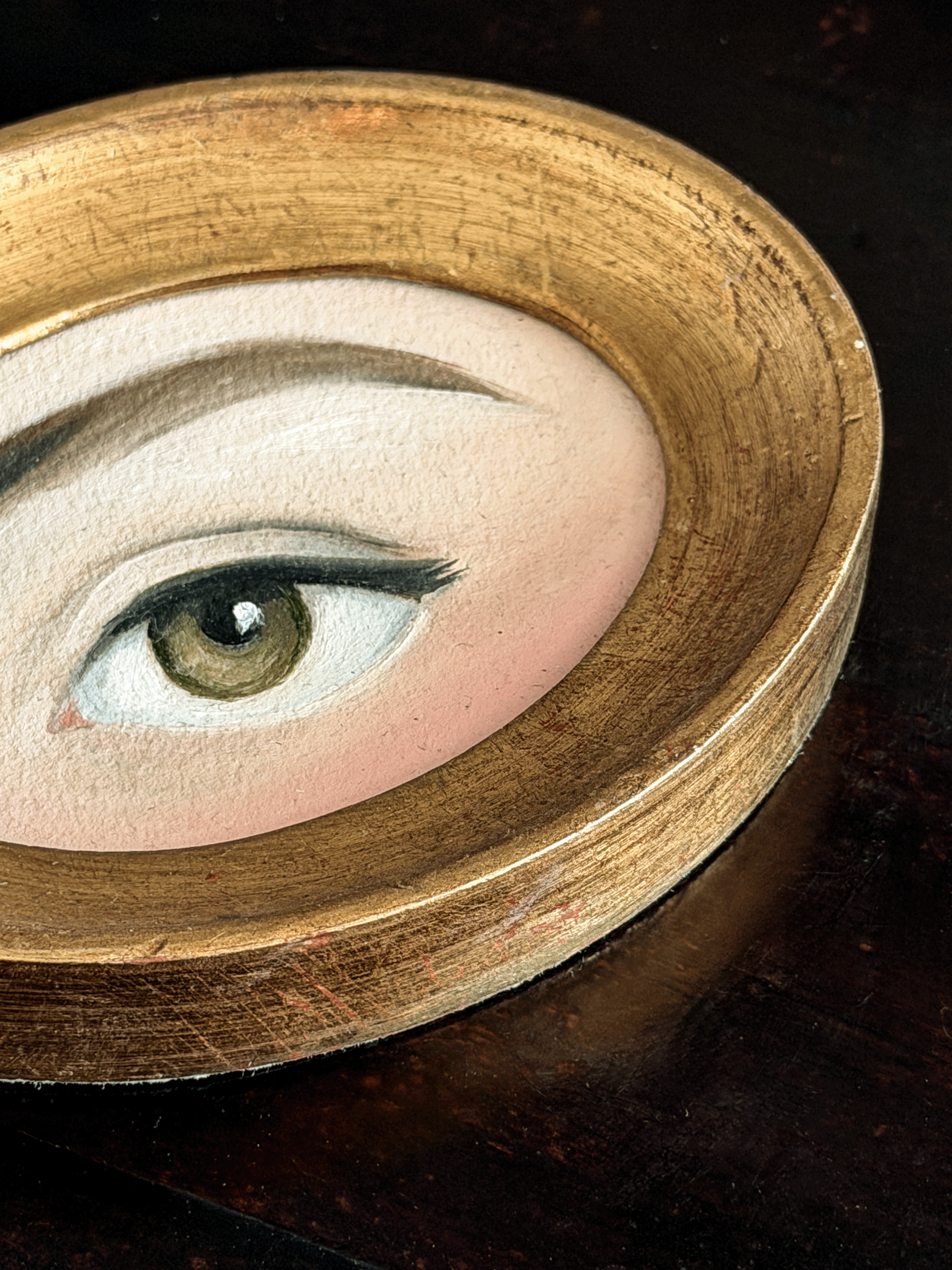A close-up of a circular wooden tray or dish holding a painted eye illustration with a realistic brown iris, black eyelashes, and pale skin tones.