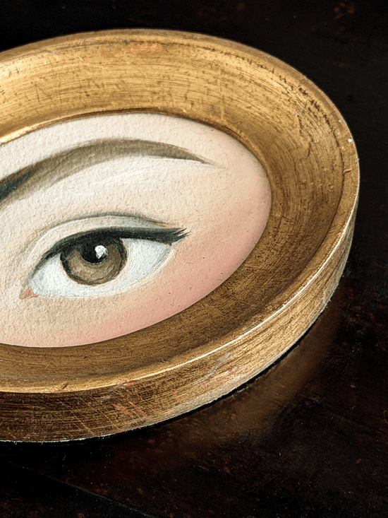 A close-up of a circular wooden tray or dish holding a painted eye illustration with a realistic brown iris, black eyelashes, and pale skin tones.