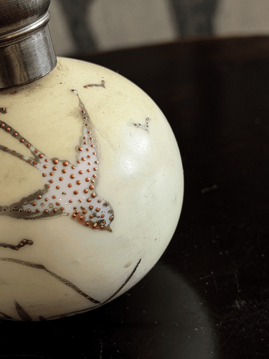 Close-up of a round ivory-colored bottle with a silver metal cap, decorated with a bird motif outlined in dark brown and filled with small metallic dots, resting on a dark surface.
