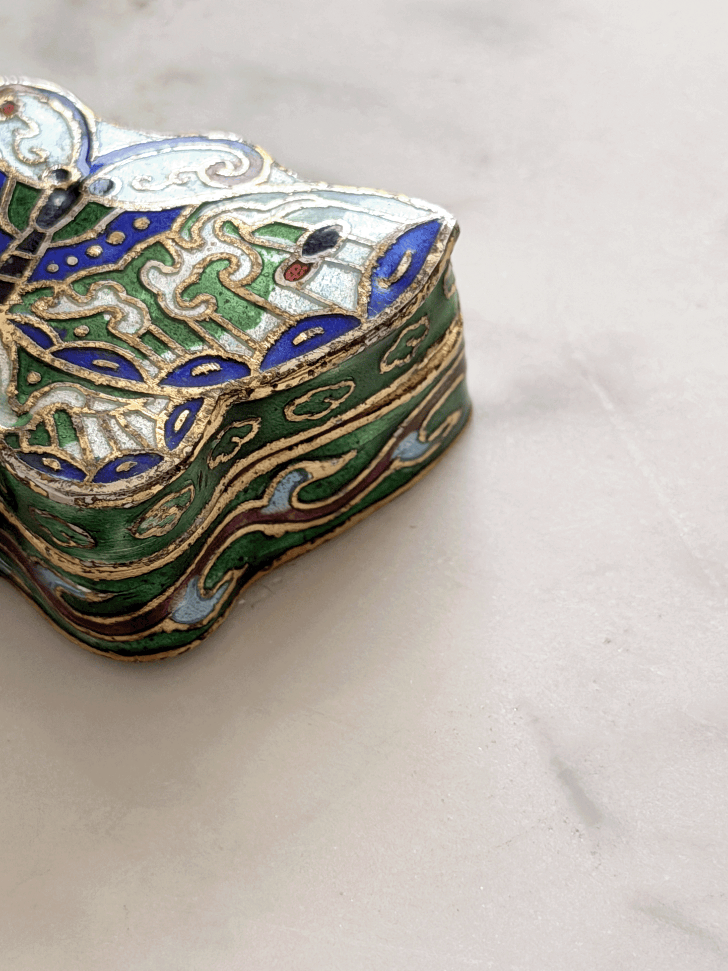 A close-up of an ornate, vintage-style enamel jewelry box with intricate green, blue, gold, and white patterns, resting on a light marble surface.