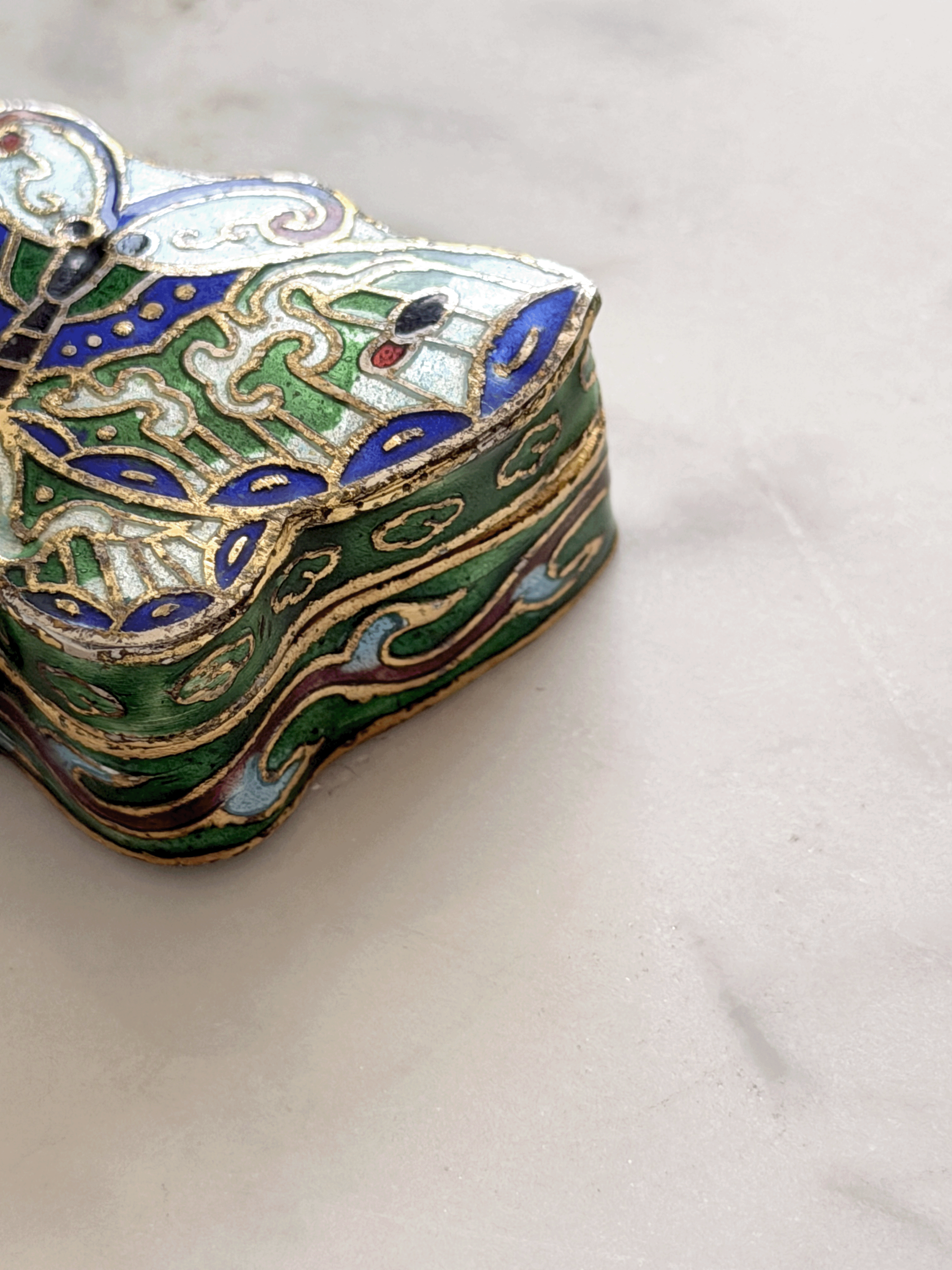 A close-up of an ornate, vintage-style enamel jewelry box with intricate green, blue, gold, and white patterns, resting on a light marble surface.