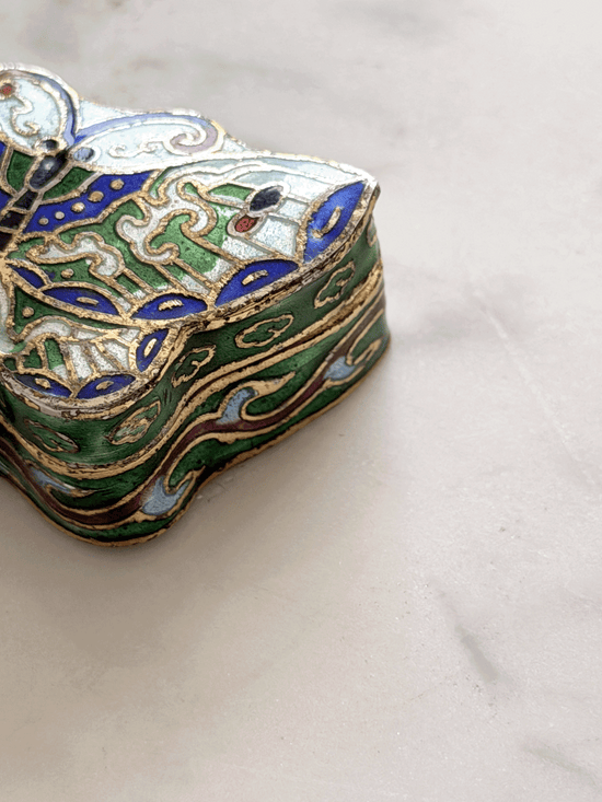 A close-up of an ornate, vintage-style enamel jewelry box with intricate green, blue, gold, and white patterns, resting on a light marble surface.