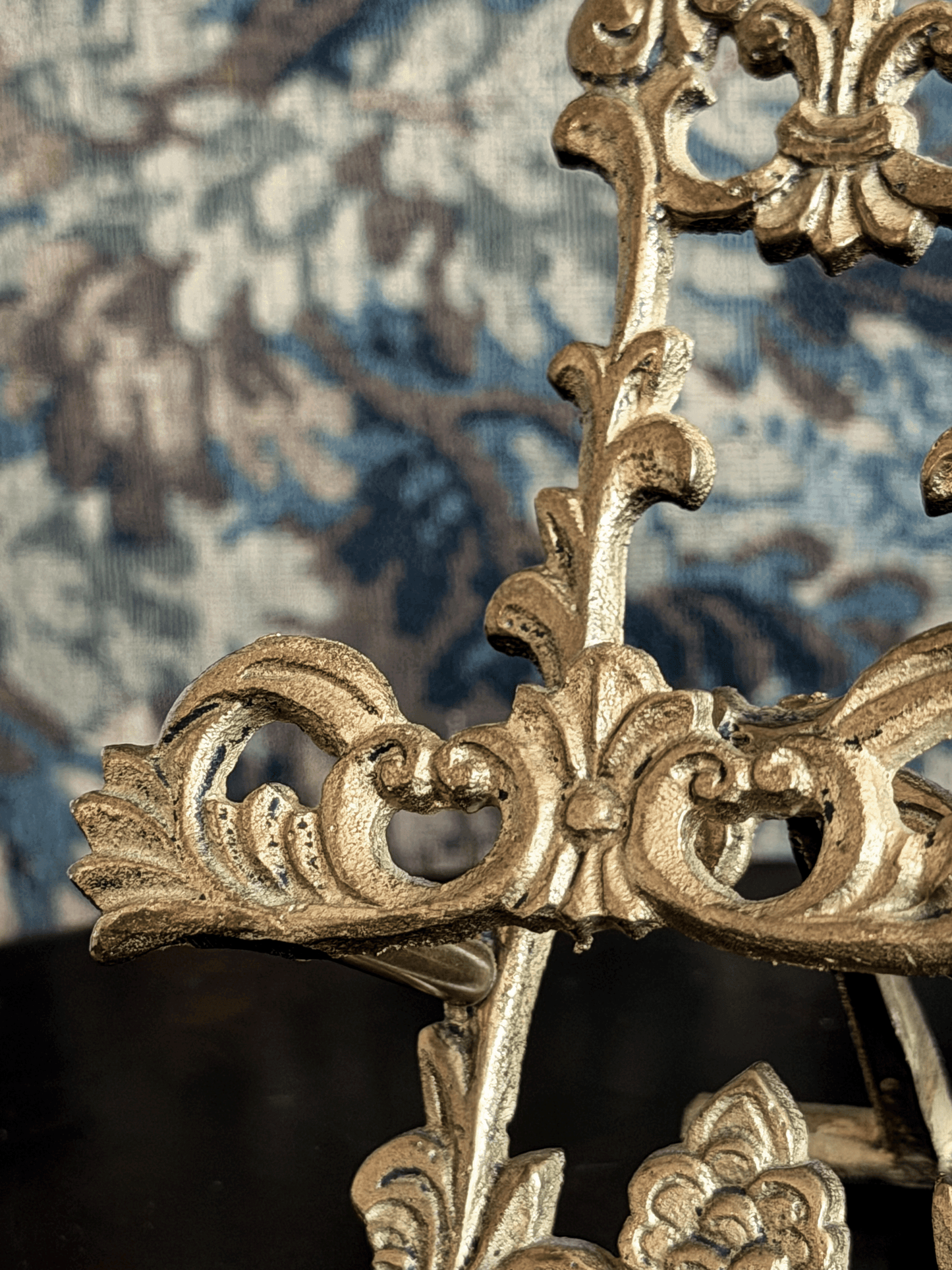 Close-up of an ornate gold metal decorative frame or chair back with carved scrollwork, set against a blue and beige patterned fabric background.