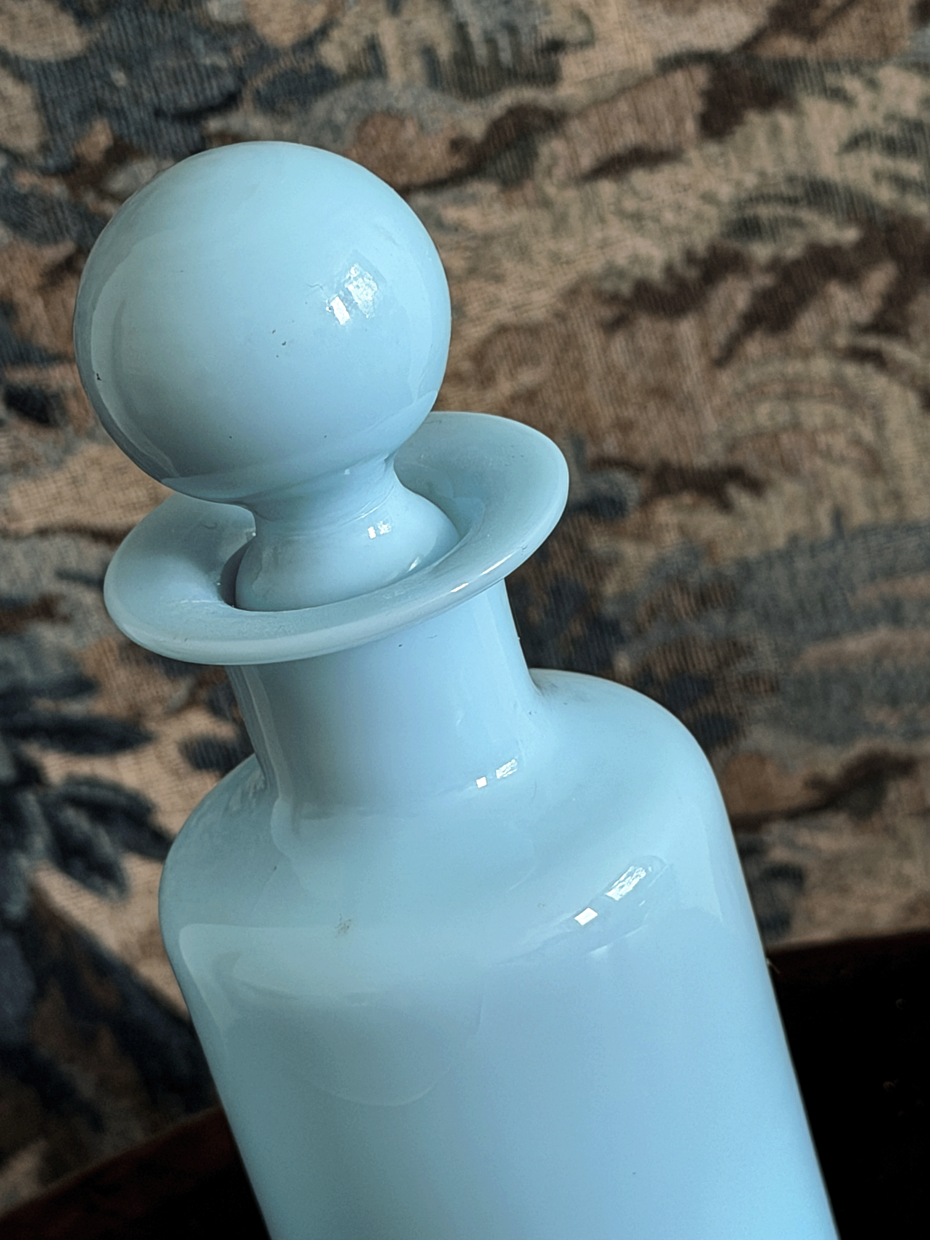 A close-up of a light blue glass opaline bottle with a matching spherical stopper, set against a dark patterned fabric background.