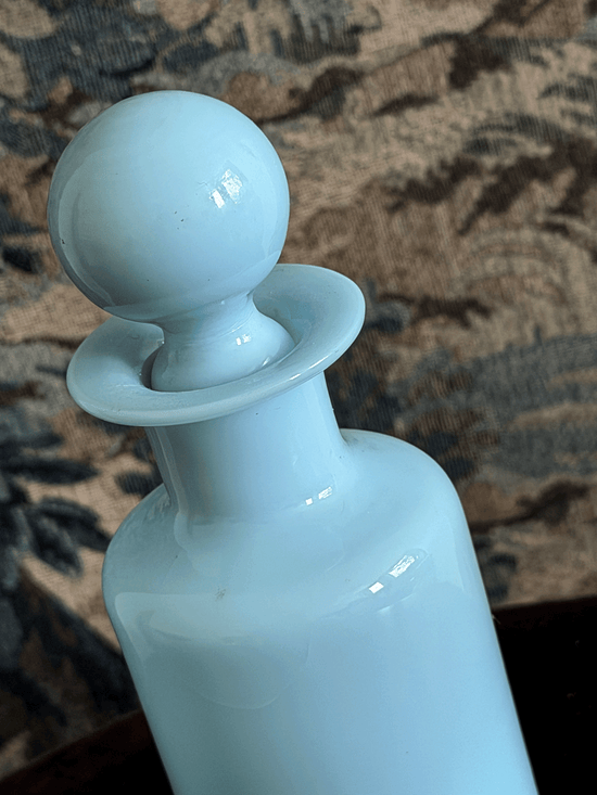 A close-up of a light blue glass opaline bottle with a matching spherical stopper, set against a dark patterned fabric background.