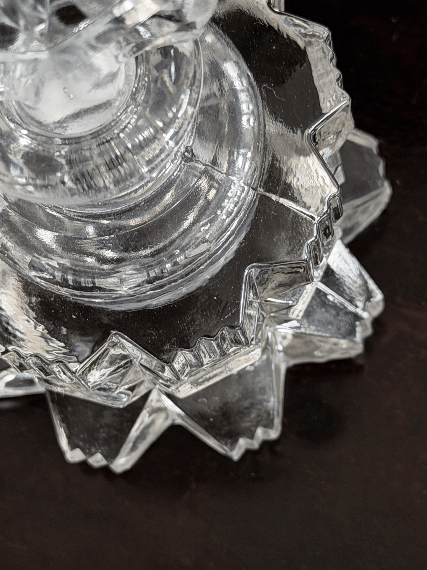 A close-up of a clear cut-glass perfume bottle with faceted, geometric edges, resting on a dark brown surface.