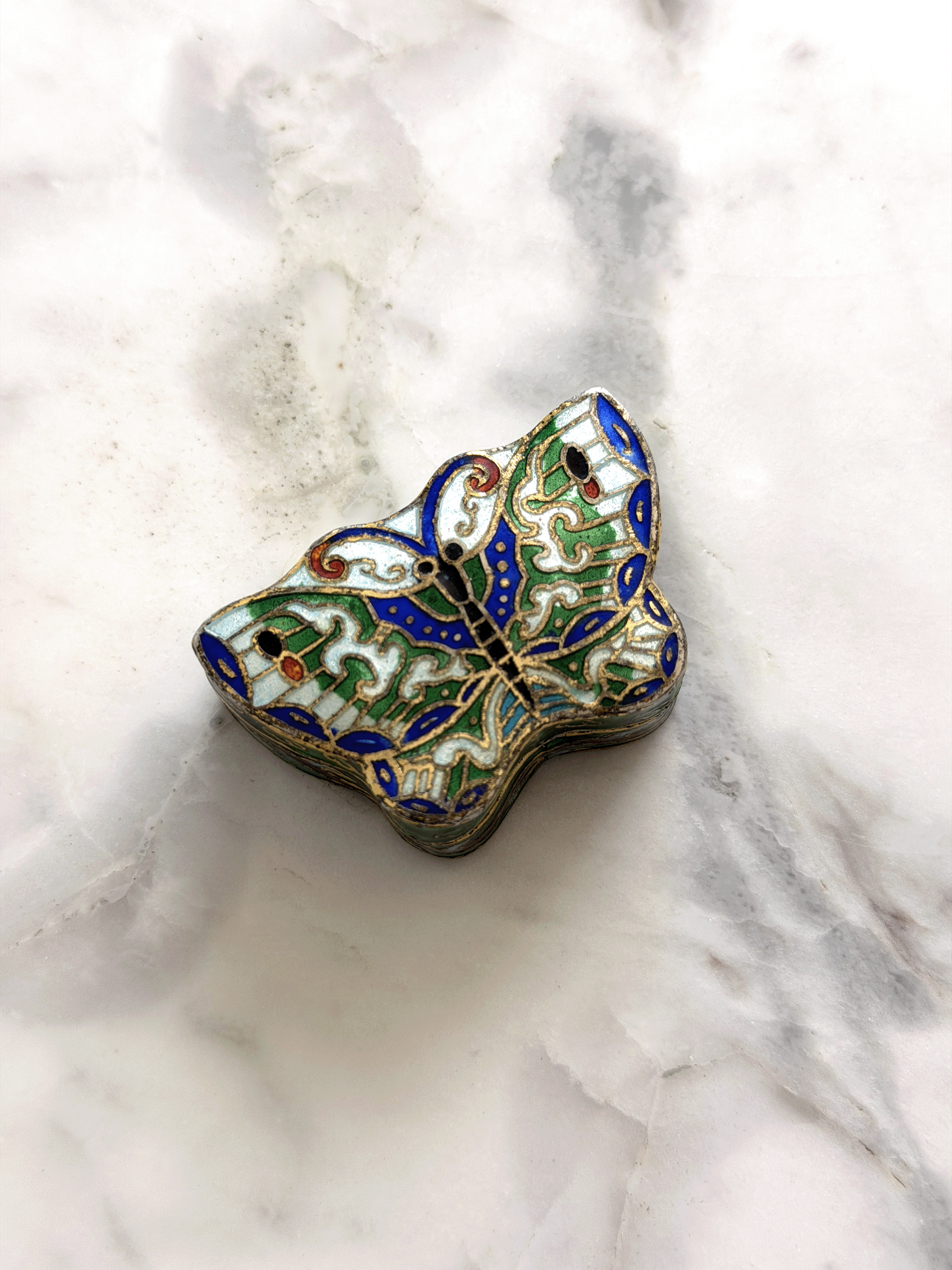 A small butterfly-shaped enamel trinket box with intricate blue, green, gold, and white detailing, resting on a white and gray marble surface.