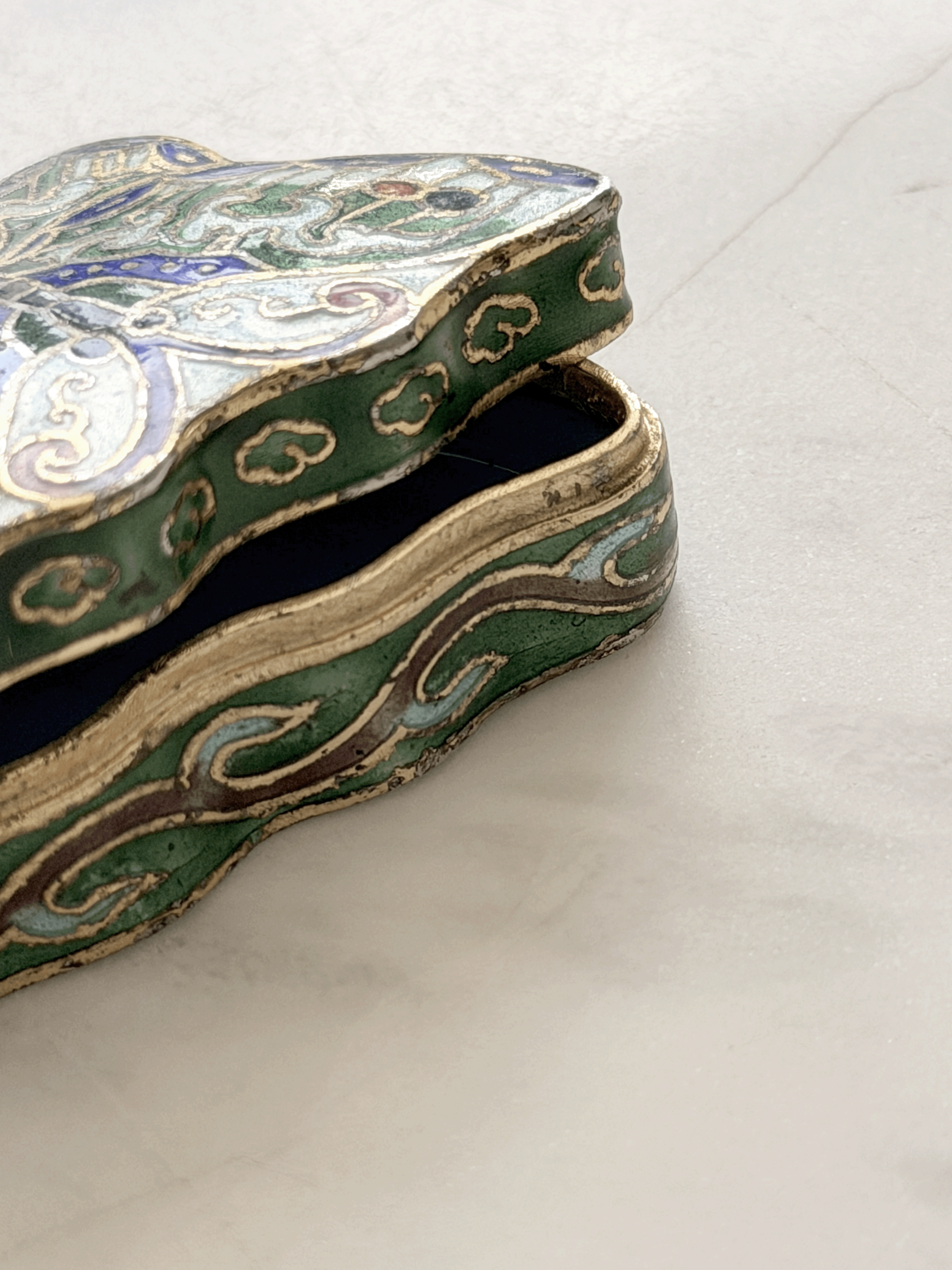 An ornate, tiny vintage butterfly-shaped trinket box with a green enamel body, gold-toned metal trim, and blue and white floral details, shown open on a light marble surface.