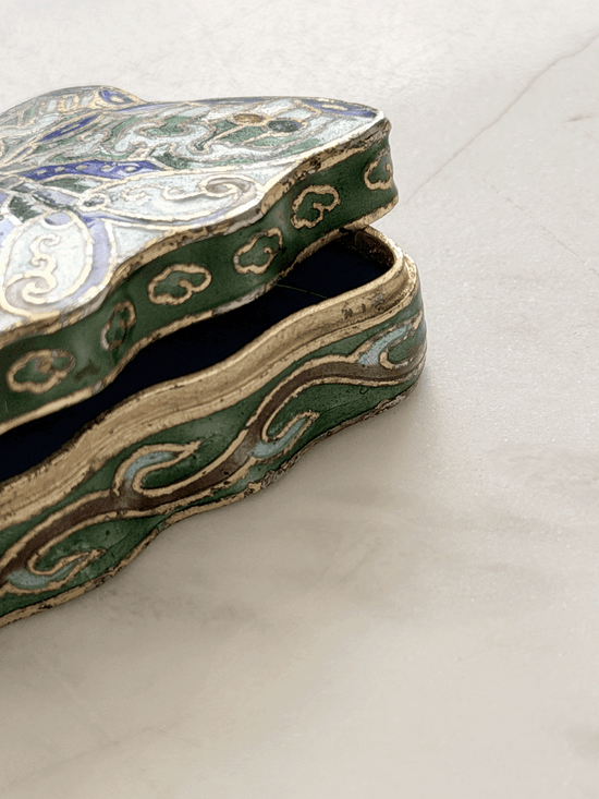 An ornate, tiny vintage butterfly-shaped trinket box with a green enamel body, gold-toned metal trim, and blue and white floral details, shown open on a light marble surface.