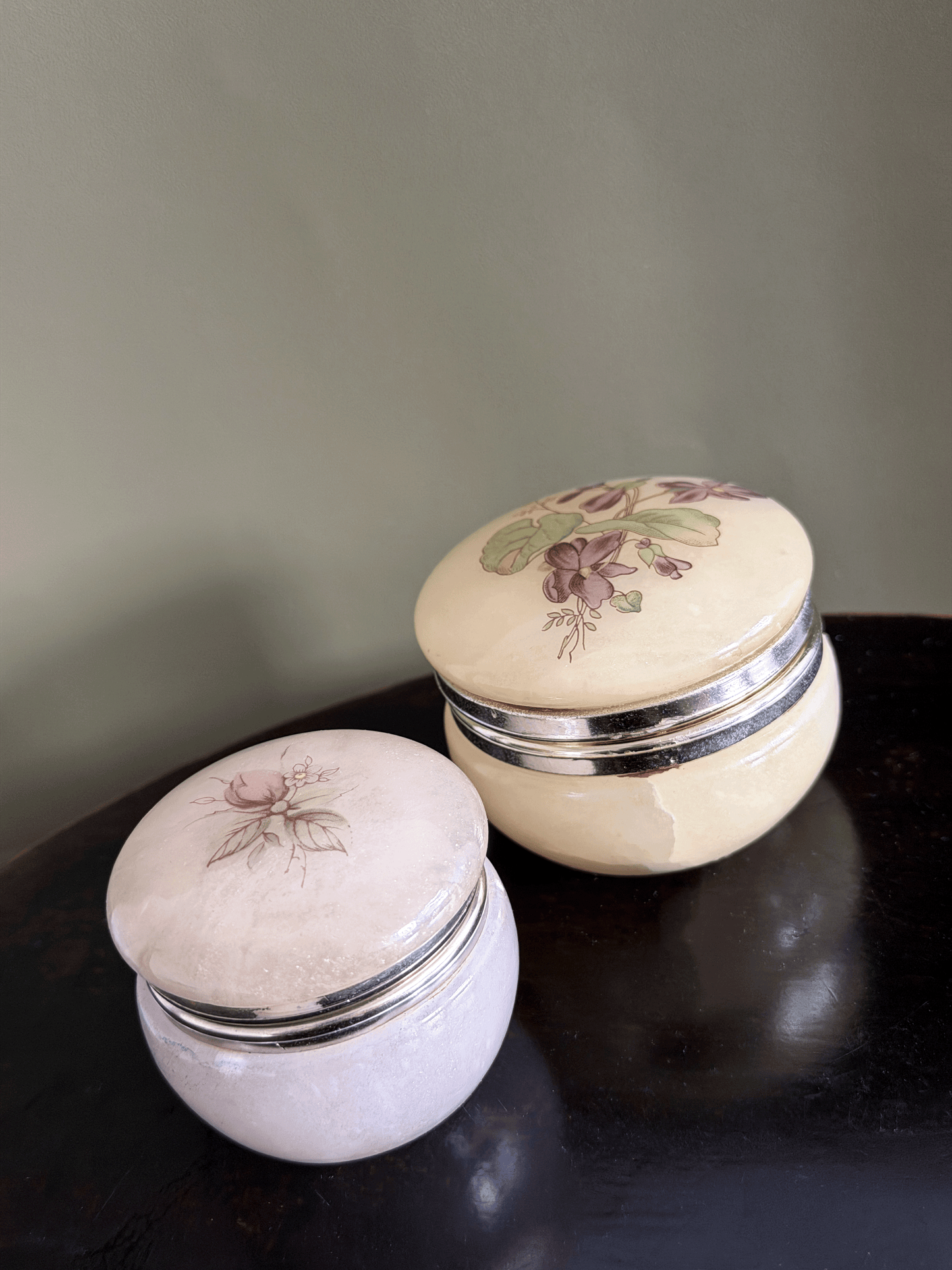 Two vintage alabaster trinket jars with gold metal bands and floral illustrations on the lids, displayed on a dark wooden surface against a muted green wall.
