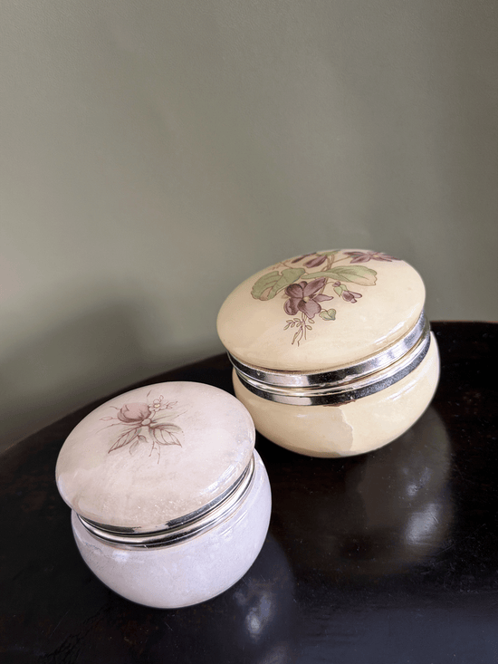 Two vintage alabaster trinket jars with gold metal bands and floral illustrations on the lids, displayed on a dark wooden surface against a muted green wall.
