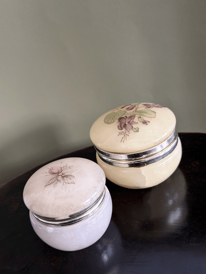 Two vintage alabaster trinket jars with gold metal bands and floral illustrations on the lids, displayed on a dark wooden surface against a muted green wall.