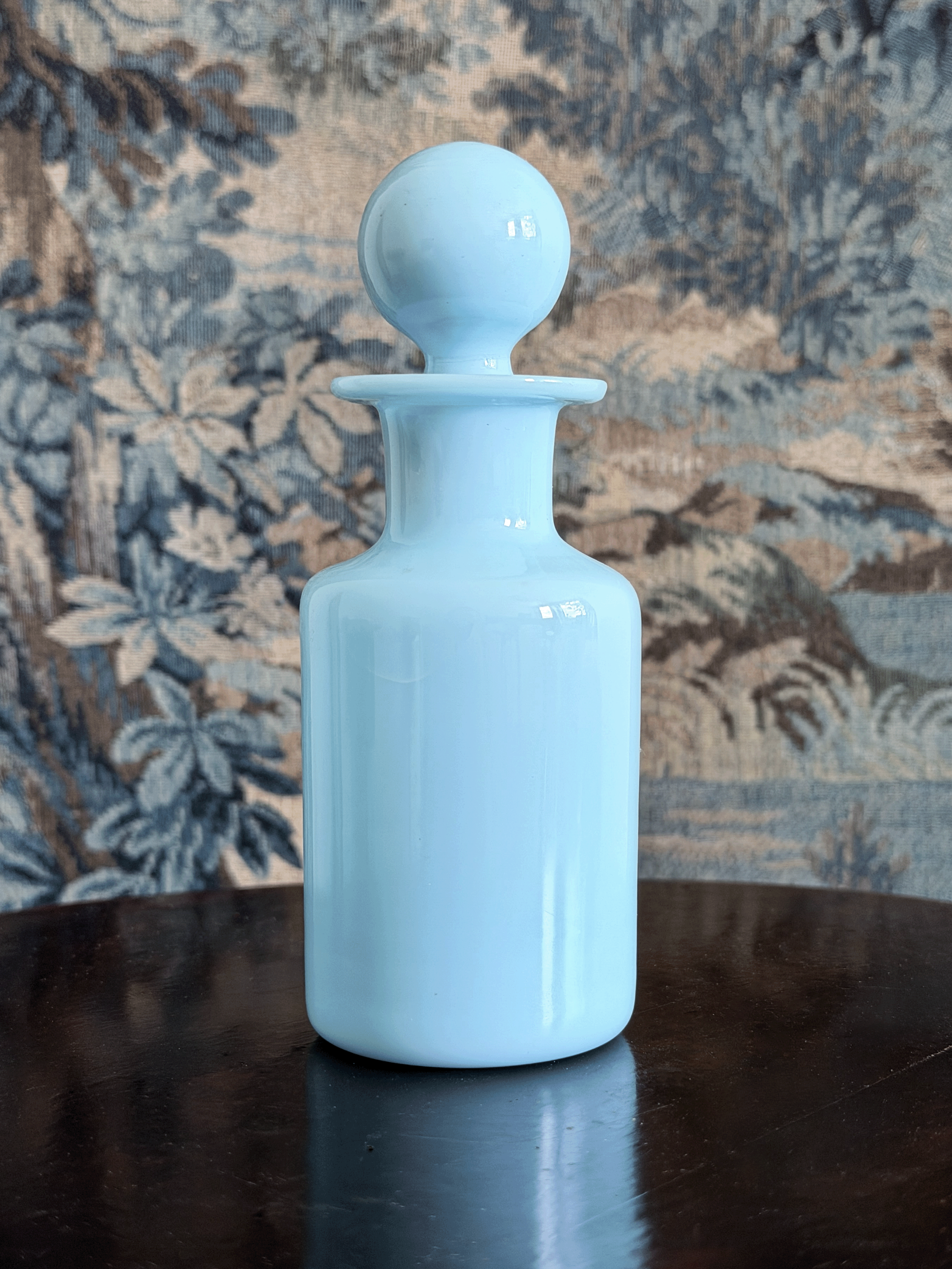 A light blue glass apothecary bottle with a matching round stopper, set on a dark reflective surface against a patterned blue and beige fabric backdrop.